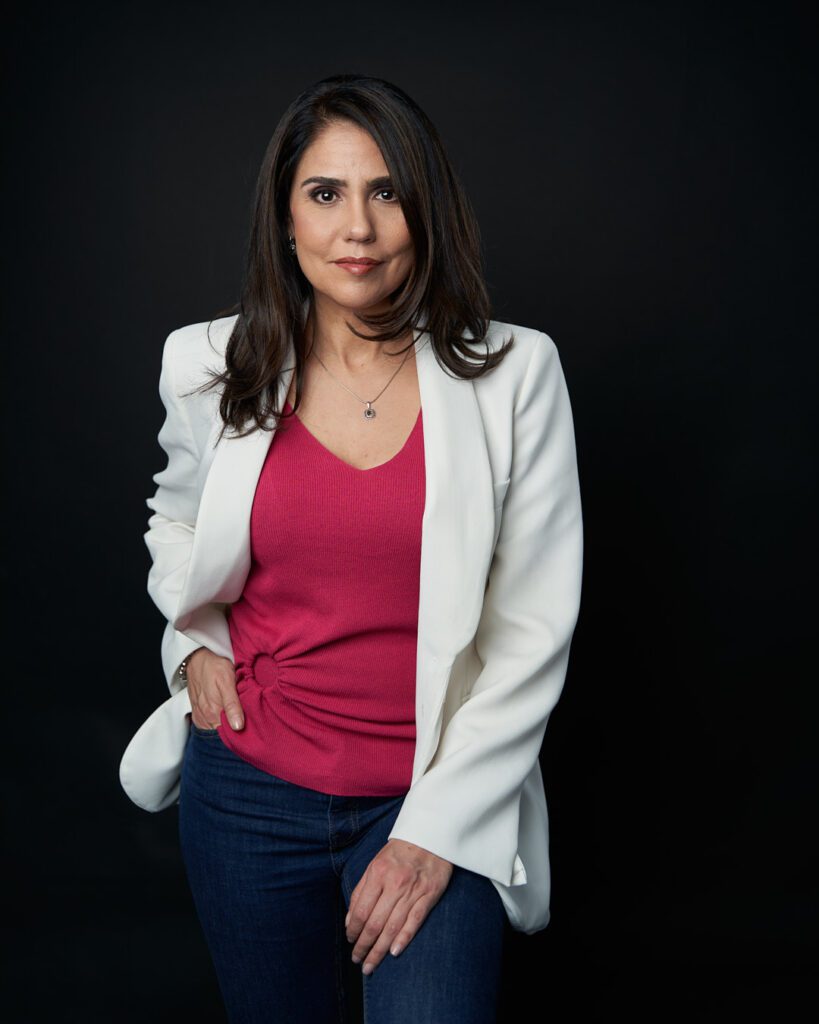 Mujer ejecutiva mostrando seguridad parada con una mano en el bolsillo de su pantalón y la otra al frente de su cuerpo, con blusa rosa y saco blanco.