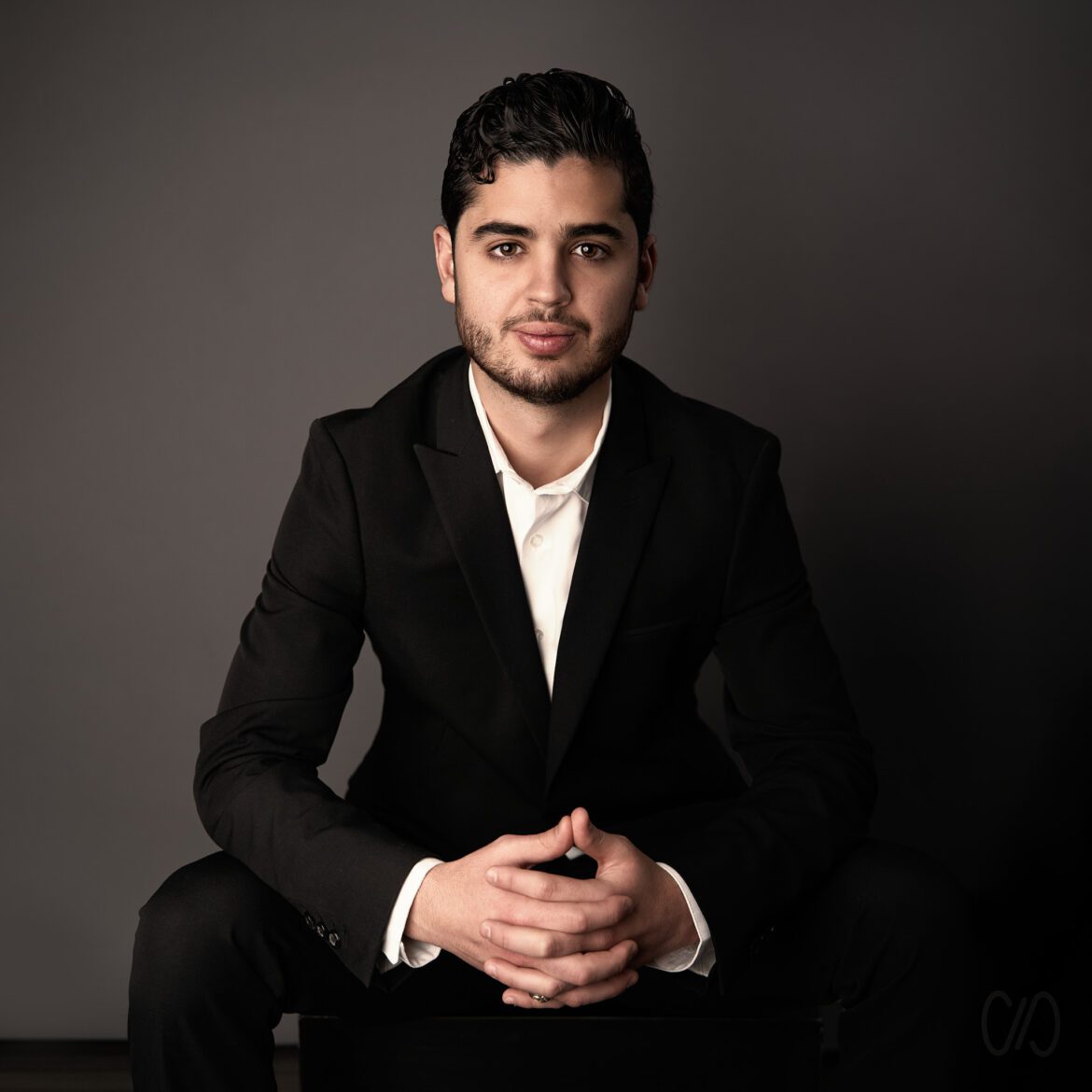 Todo sobre Retrato Fotográfico mostrando a un joven sentado recargado sobre sus brazos con sus manos entrelazadas vistiendo traje ejecutivo