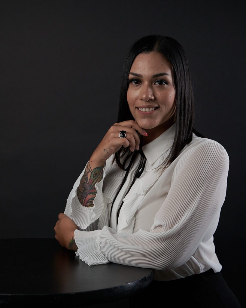 Mujer en foto de perfil con la mirada hacia la cámara con blusa blanca y recargada en una mesa