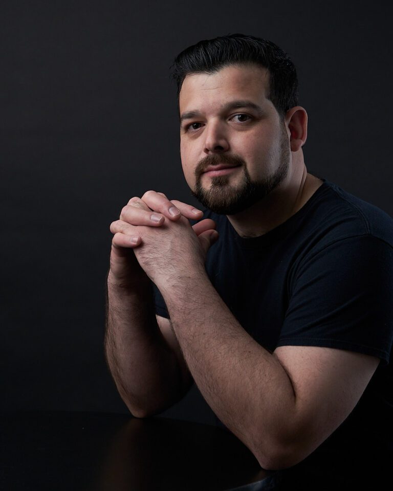 Hombre con barba y playera negra sobre fondo negro con los brazos apoyados sobre una mesa y el mentón echado hacia atrás