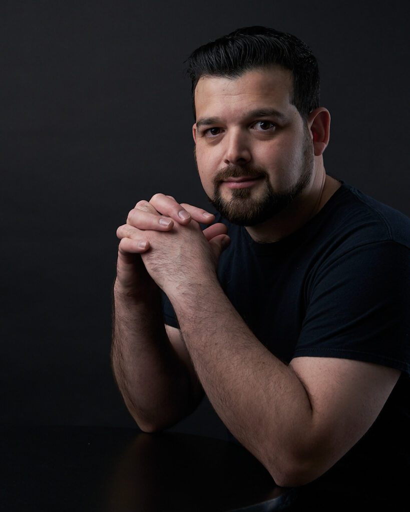 Hombre con barba y playera negra sobre fondo negro con los brazos apoyados sobre una mesa