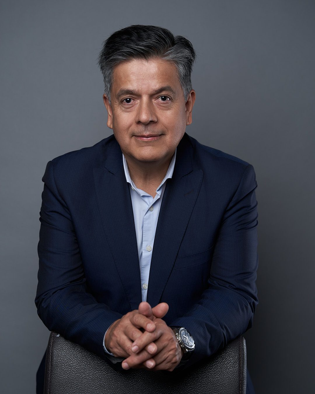 Foto de perfil ejecutivo de hombre maduro con revelado y edición profesional de piel, usando saco azul recargado sobre un banco con las manos juntas