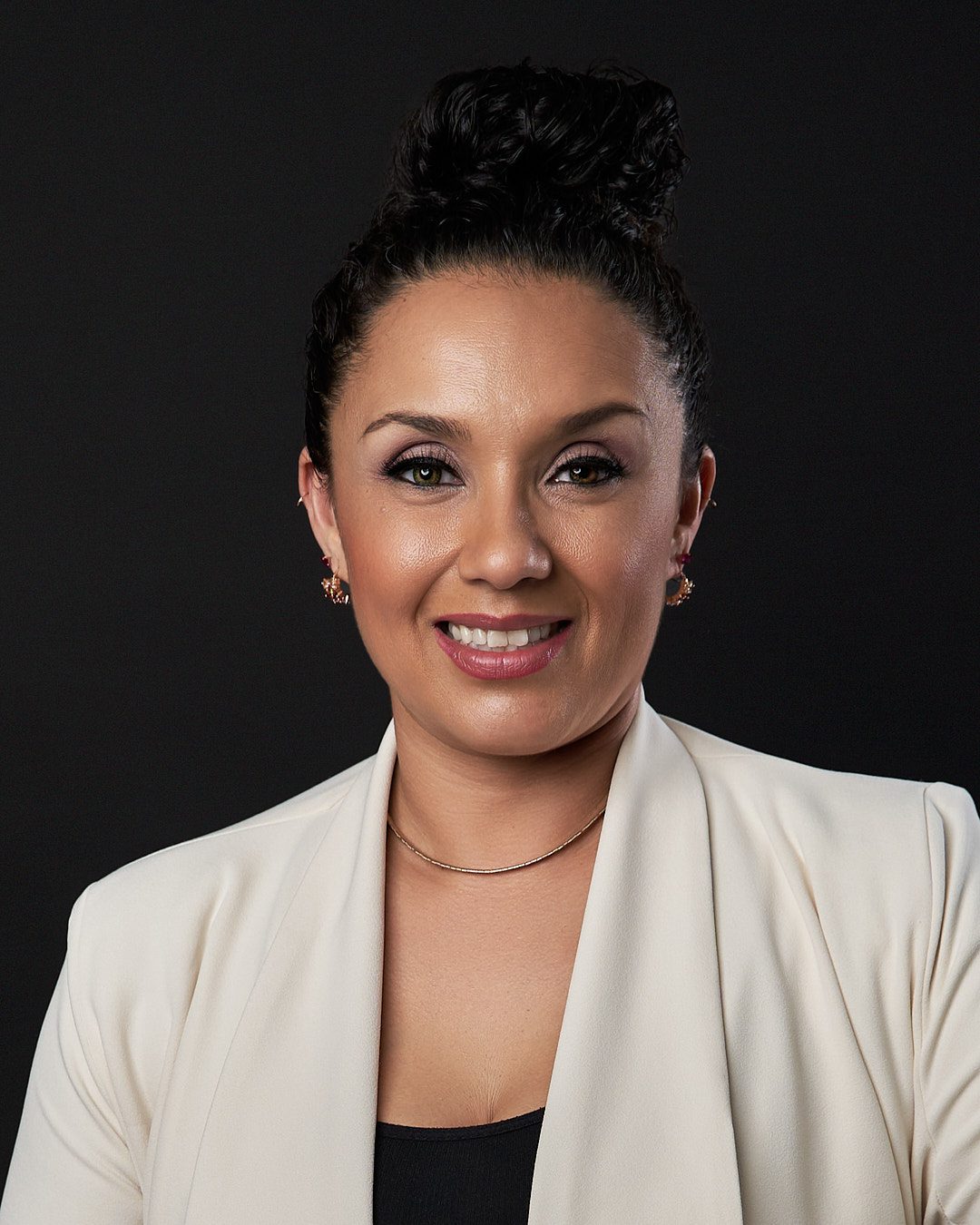Headshot de mujer con cabello rizado, en cebolla arriba de la cabeza y con blusa negra con saco blanco sonriendo luciendo un retoque digital en su rostro mejorando el maquillaje