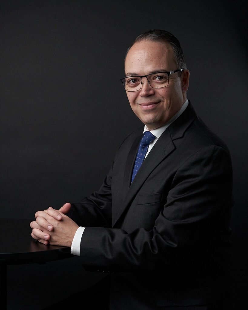 Hombre de frente ancha con lentes y expresión sonriente visitiendo traje gris y corbata azul con las manos entrelazadas y apoyadas sobre una mesa me ayuda a explicar las 10 Reglas de Etiqueta en los Negocios Alrededor de Mundo