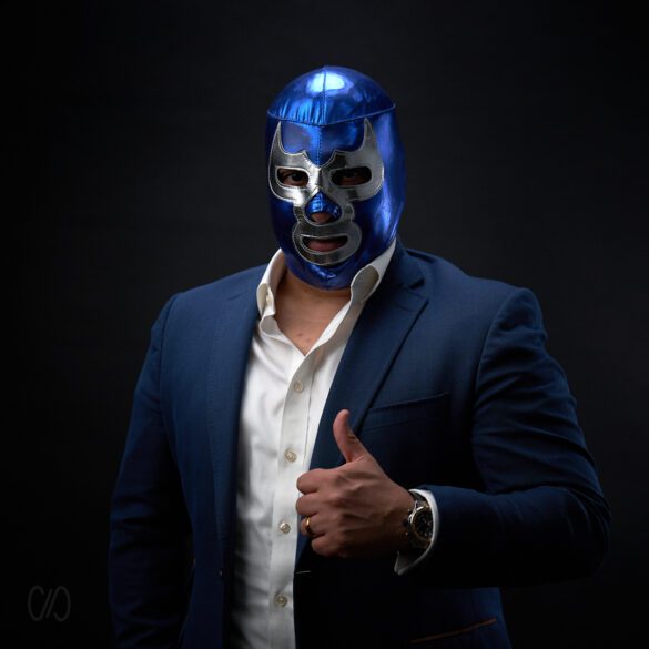 hombre con camisa de vestir y saco azul con mascara en la cara de luchador en tono azul y plateada con la mano levantando el dedo pulgar